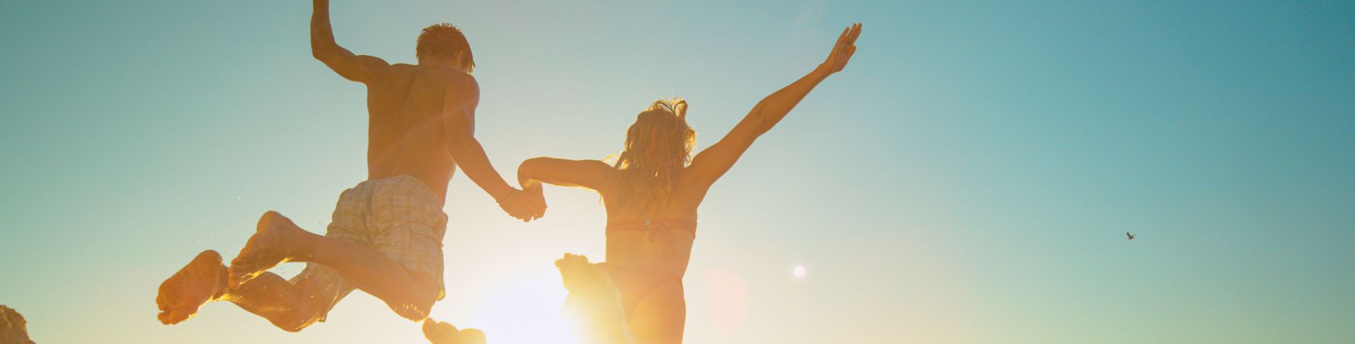 The sun shines on a couple leaping off a cliff into the water below.