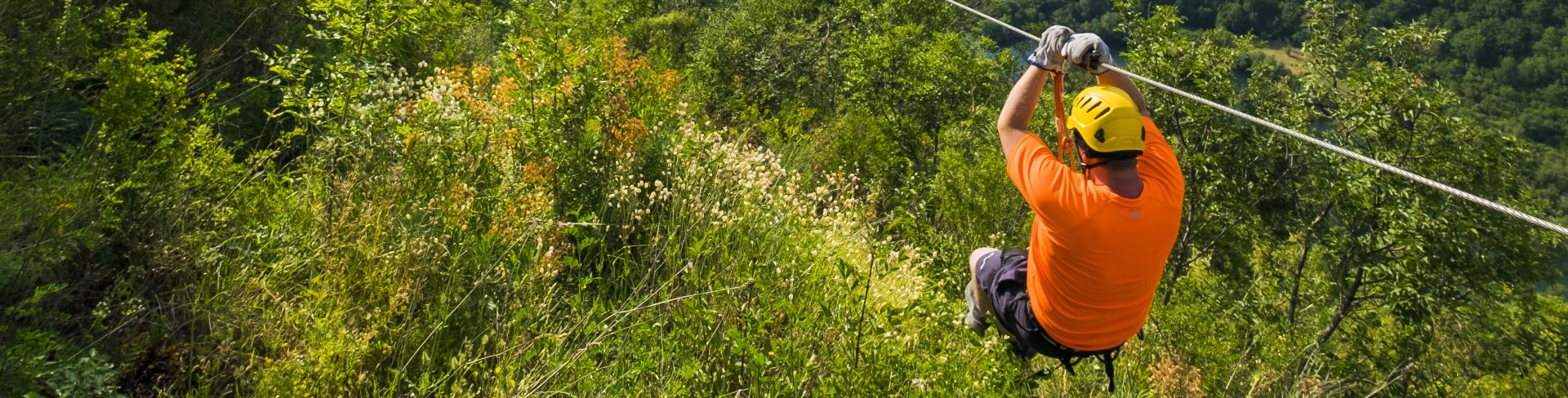 A man in an orange shirt holds on as he zip-lines through the green forest.