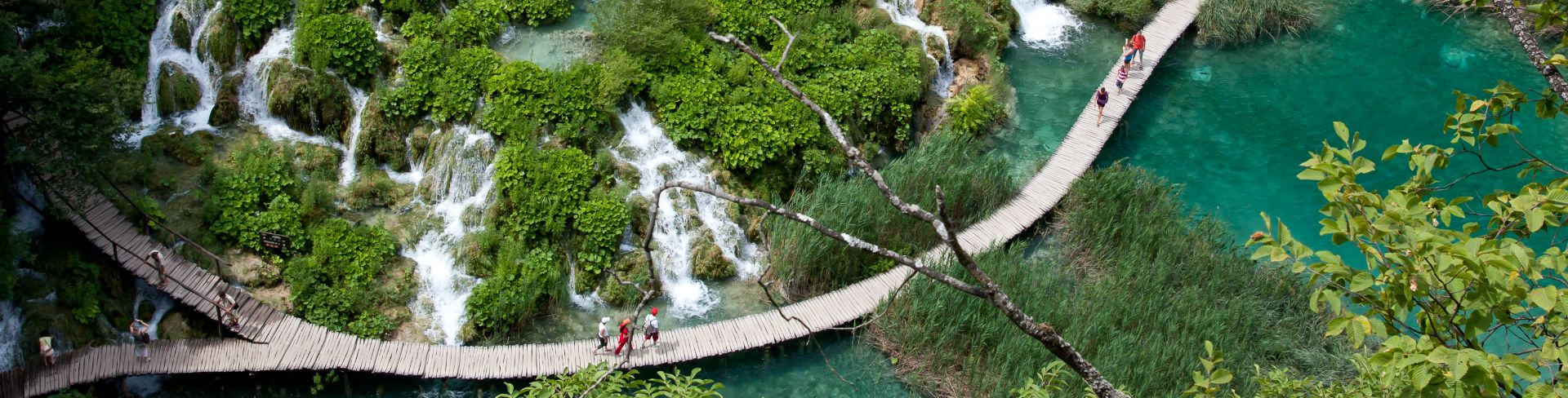 People walk along a wooden pathway by waterfalls and green plant life. 