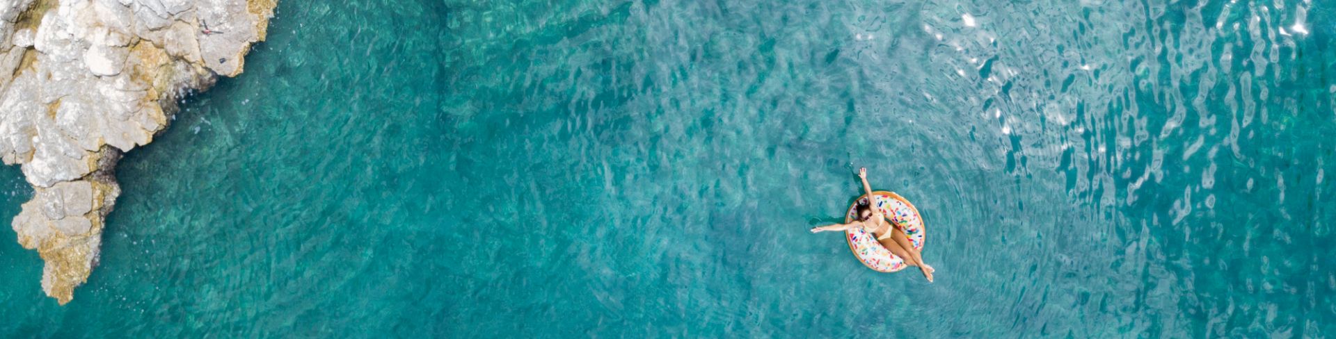 A woman floats in the blue waters beside a rock cliff.