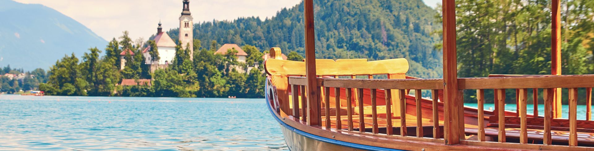 A wooden boat floats toward a bell tower nestled in green hills in the distance. 