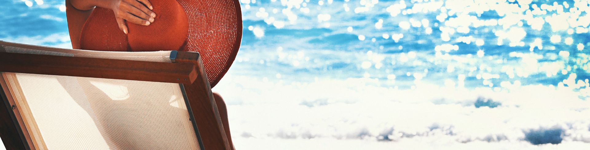 The rear view of a woman wearing a large straw hat and reclining on a sun lounger on the beach.