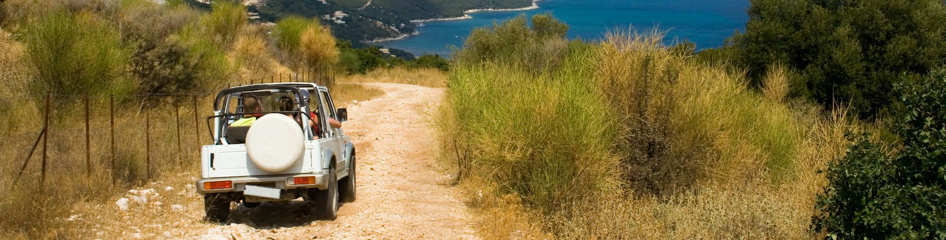 The rear view of a white Jeep with the top down, driving on a rocky path with views of the sea.