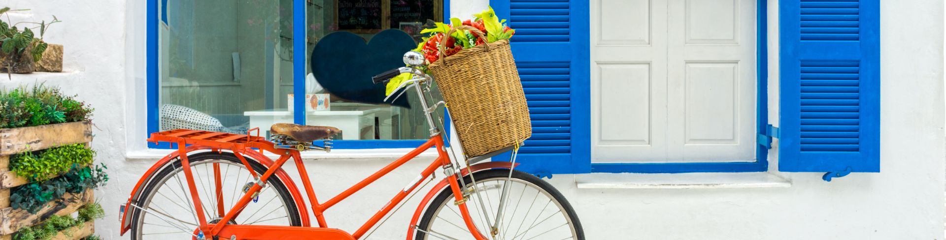 An orange bicycle with a basket of flowers parked in front of a whitewashed building with blue shutters.