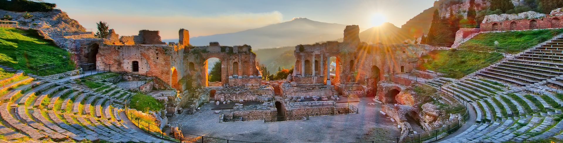 The remains of an ancient amphitheater with Mount Etna in the background at sunset.