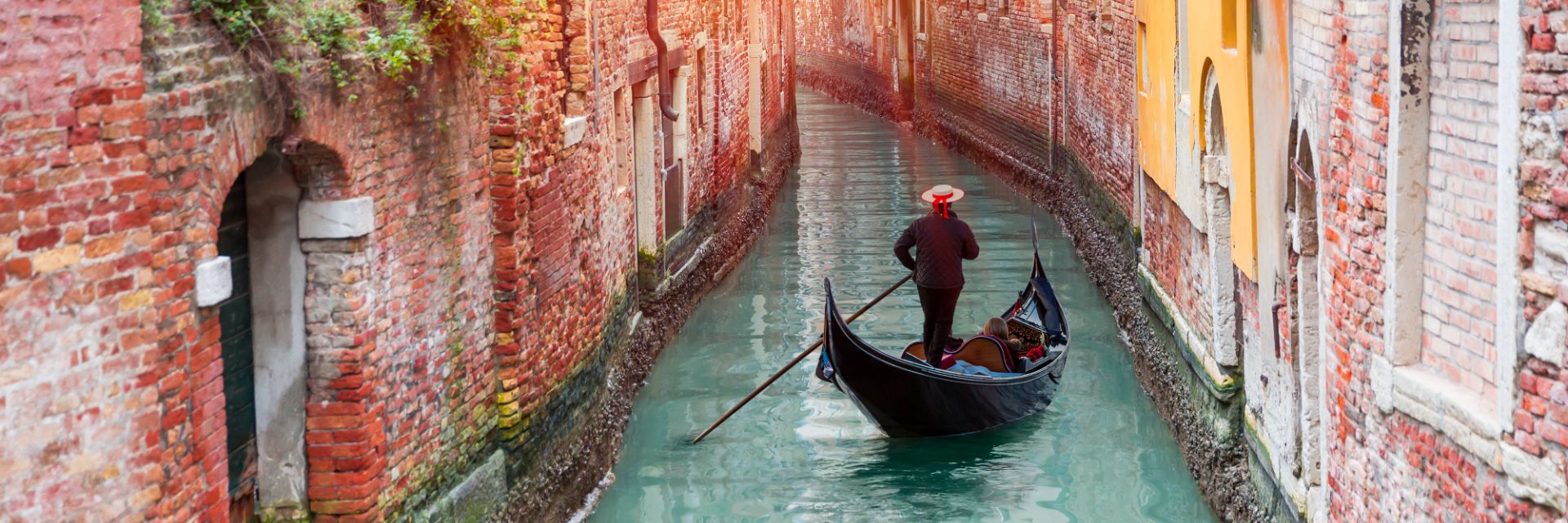 A gondola is steered through a narrow waterway between two red brick walls.
