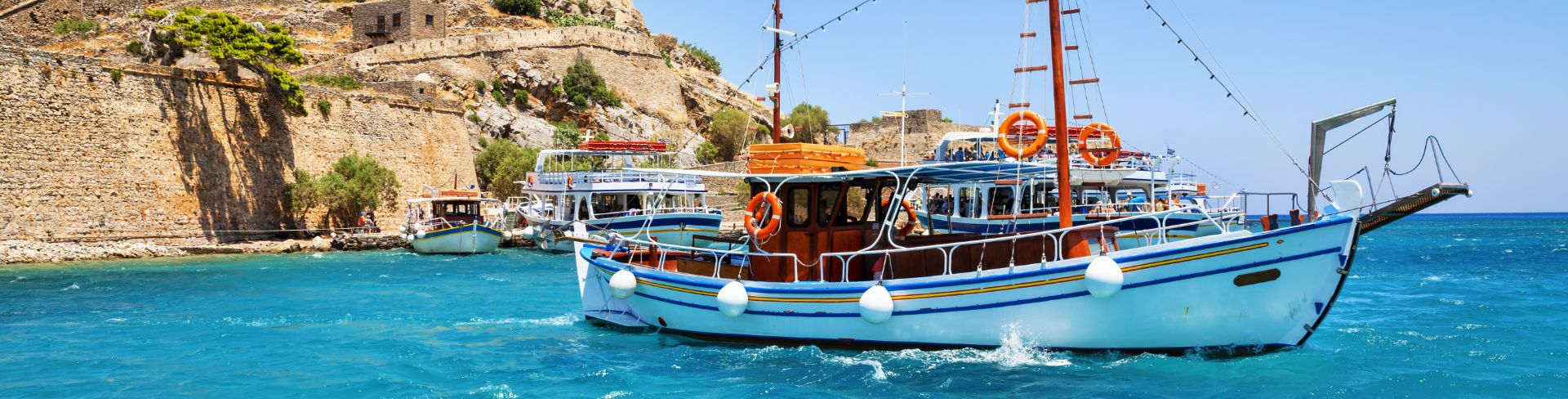 A fishing boat on clear blue Mediterranean waters with Spinalonga island in the background.