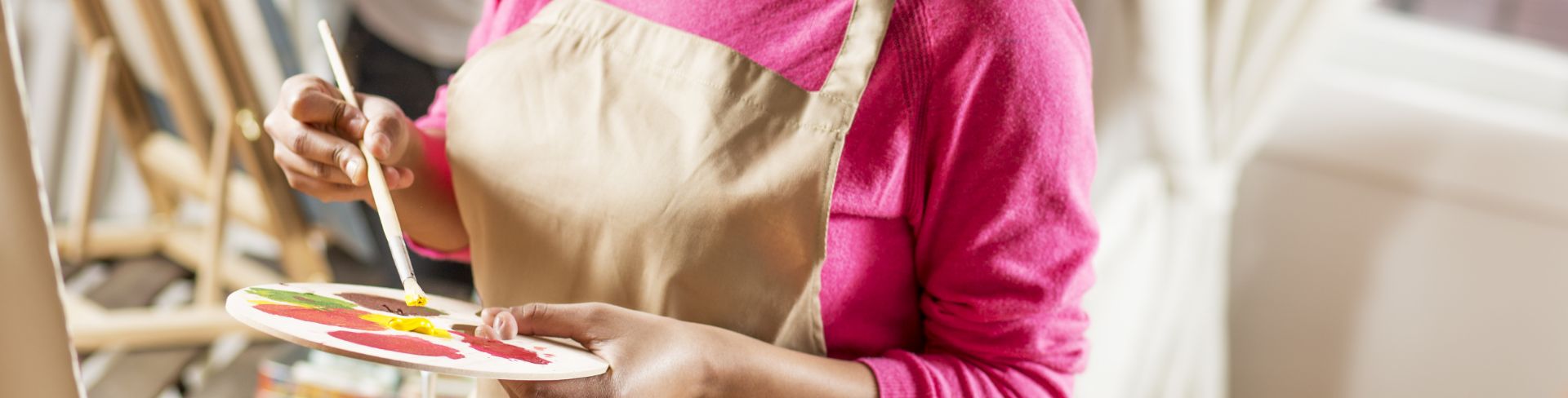 A hand paints on a canvas while the other hand holds a watercolor palette full of different colors.
