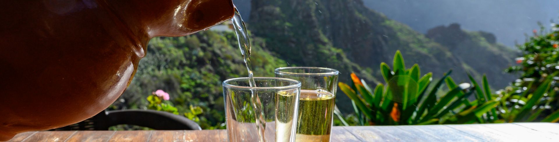 A clay pouring white wine into a glass on a terrace with lush mountains in the background.