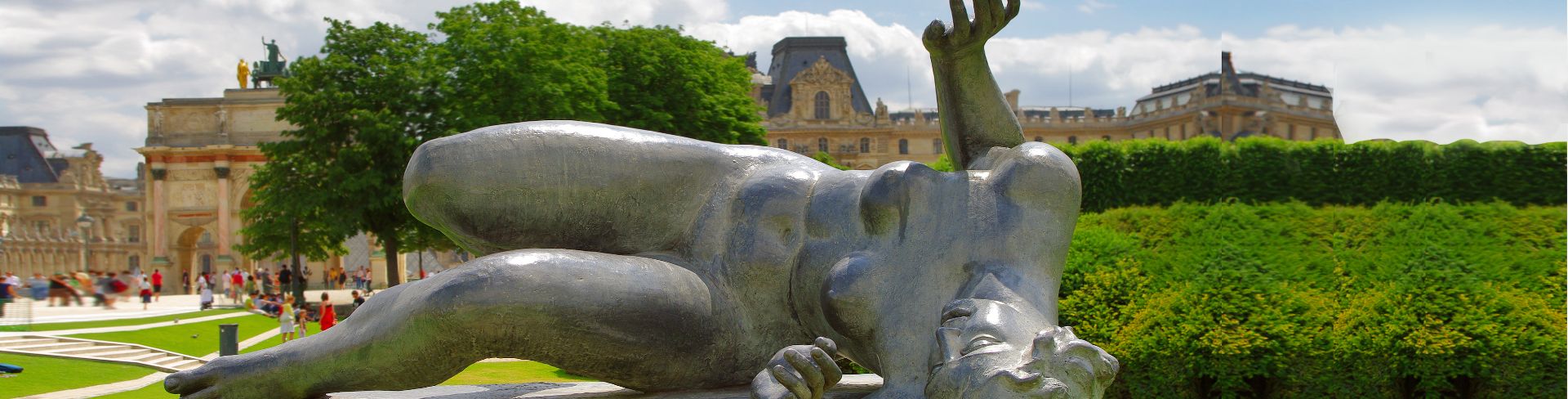 A silver sculpture of a woman lying on her side in a green park with a historical building in the background.
