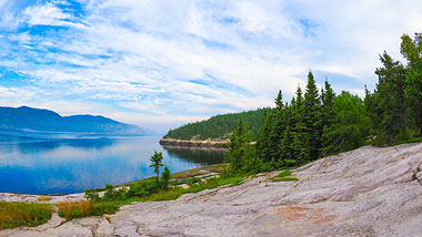 White clouds streak the blue sky over deep blue water, and a rock shoreline and green trees.