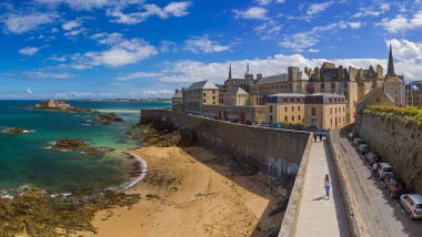 Blue green water in front of a beach where a great wall leads to an old castle.
