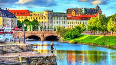 Colorful buildings and bright green grass line the bridge leading to water.