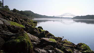 Dark rocks covered in green moss line the banks of the Cape Cod Canal, a dark canal with a bridge in the distance.