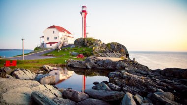 A tall red and white light house sits next to a house on a grassy point in front of the water.