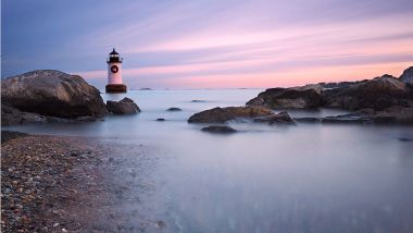 A white lighthouse sits at the rocky shore's edge as sun sets.