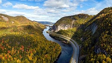 Mountains covered in green and yellow trees, through which a river and road run.